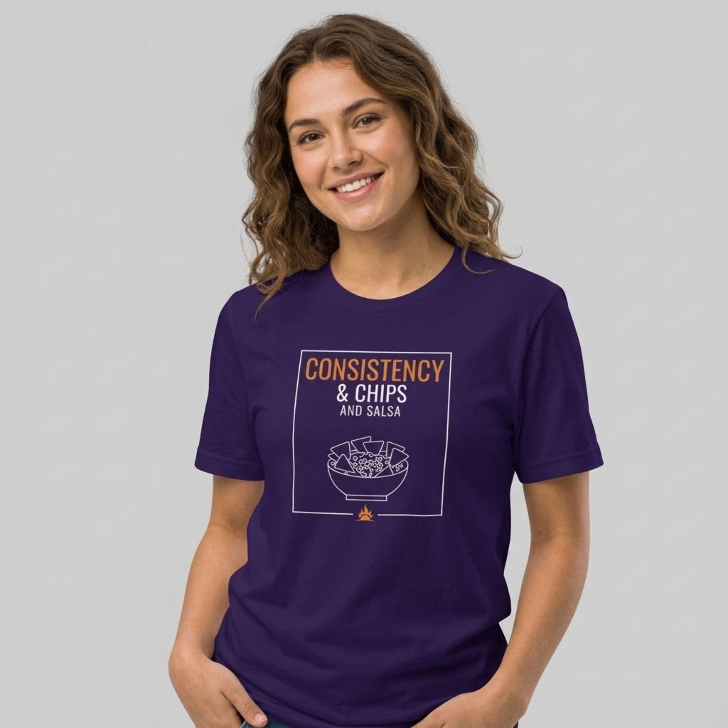 A woman wearing a dark purple short-sleeve t-shirt with a graphic print that reads 'CONSISTENCY & CHIPS AND SALSA' above a bowl illustration, photographed against a solid light gray background.