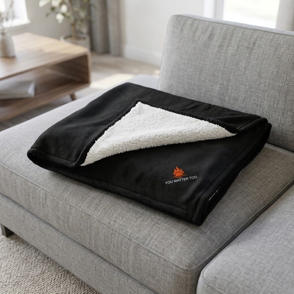 A black fleece throw blanket with a white sherpa lining is folded and placed on a light gray fabric sofa. The front of the blanket is black with a small orange campfire logo and the words 'YOU MATTER TOO' printed near the bottom corner.
