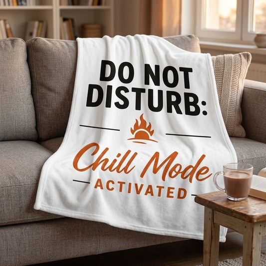 A white fleece throw blanket draped over a gray couch in a cozy living room setting. The blanket features the text 'DO NOT DISTURB:' in black, with an orange flame icon, and 'Chill Mode ACTIVATED' in orange script and block lettering.