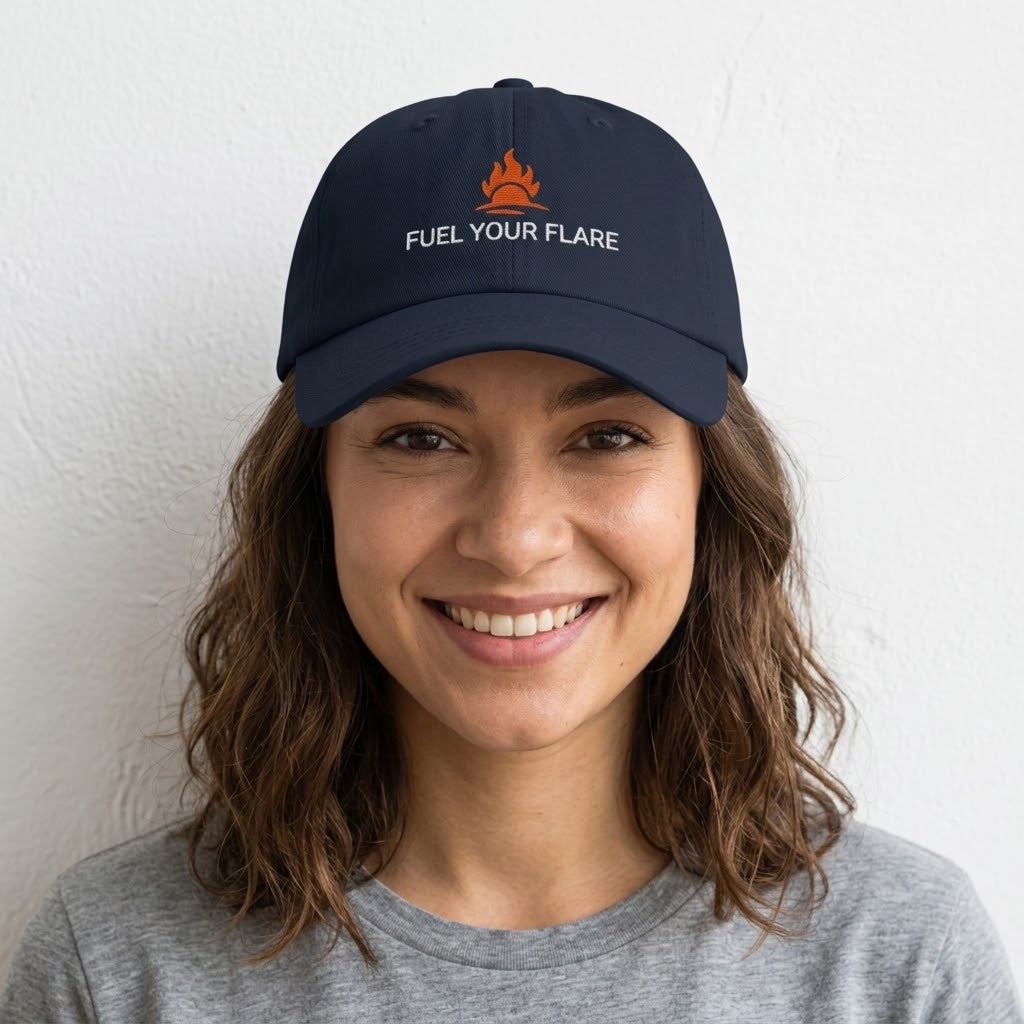 A smiling woman with wavy brown hair wearing a navy blue baseball cap with an orange campfire logo and the white embroidered text 'FUEL YOUR FLARE' against a plain white wall.