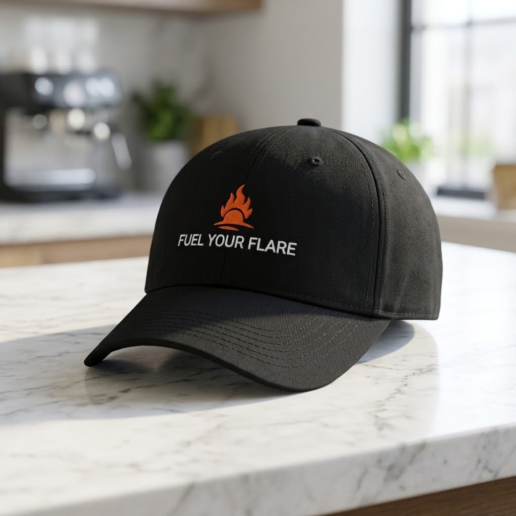 A black baseball cap with an embroidered orange flame logo and the white text 'FUEL YOUR FLARE' on the front, resting on a white marble countertop in a blurred kitchen setting.
