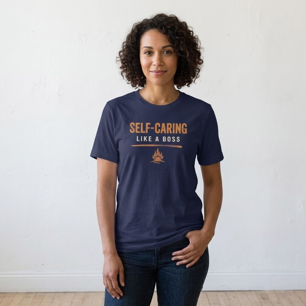 A woman with curly dark hair stands against a white wall wearing a navy blue graphic t-shirt with the text 'SELF-CARING LIKE A BOSS' in orange and a small campfire graphic. She is also wearing dark blue jeans.