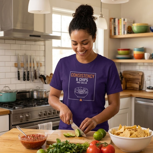 A woman in a kitchen wearing a purple graphic t-shirt that reads 'CONSISTENCY & CHIPS AND SALSA'. She is slicing lime on a wooden cutting board next to a bowl of salsa and chips.