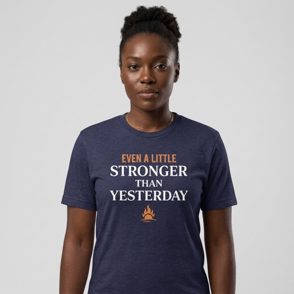 A woman wearing a navy blue heathered t-shirt with the text 'EVEN A LITTLE STRONGER THAN YESTERDAY' in white and orange, along with a small campfire graphic. She is standing against a neutral light gray background.