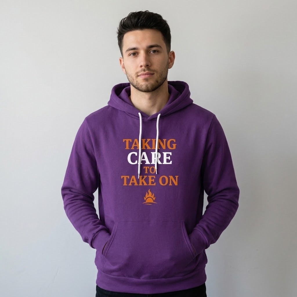 A man wearing a purple pullover hoodie with the text 'TAKING CARE TO TAKE ON' in orange and white, paired with black pants, standing against a plain light gray wall.