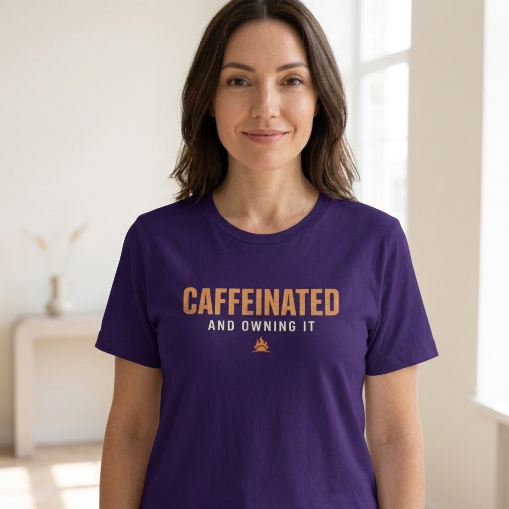 A woman wearing a team purple short-sleeve t-shirt with the text 'CAFFEINATED AND OWING IT' in orange and white, standing in a brightly lit indoor room.