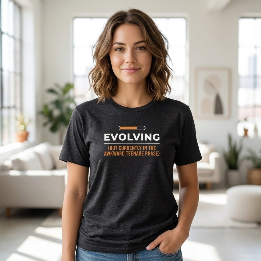 A woman wearing a dark heather gray graphic t-shirt with the text 'EVOLVING (BUT CURRENTLY IN THE AWKWARD TEENAGE PHASE)' in white and orange, standing in a bright, modern living room.