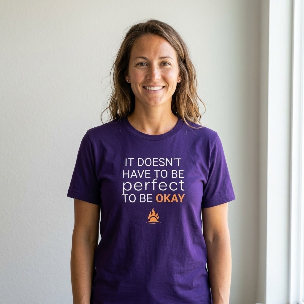 A smiling woman with light brown hair stands indoors wearing a purple short-sleeve T-shirt with the text 'IT DOESN'T HAVE TO BE perfect TO BE OKAY' in white and orange lettering, plus a small orange flare graphic below.