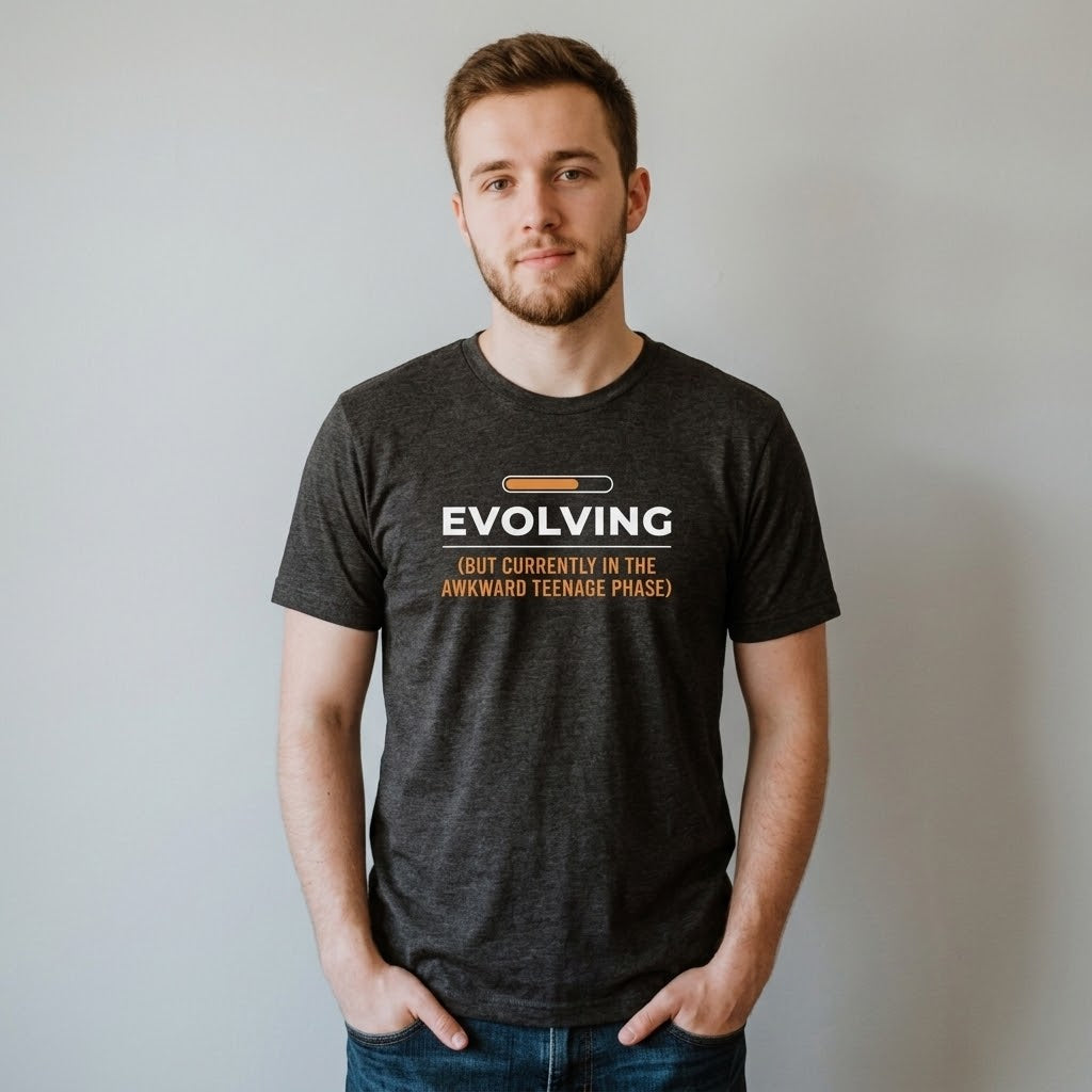 A man wearing a dark heather gray t-shirt with a graphic that reads 'EVOLVING (BUT CURRENTLY IN THE AWKWARD TEENAGE PHASE)' in white and orange text, standing against a plain light gray wall.